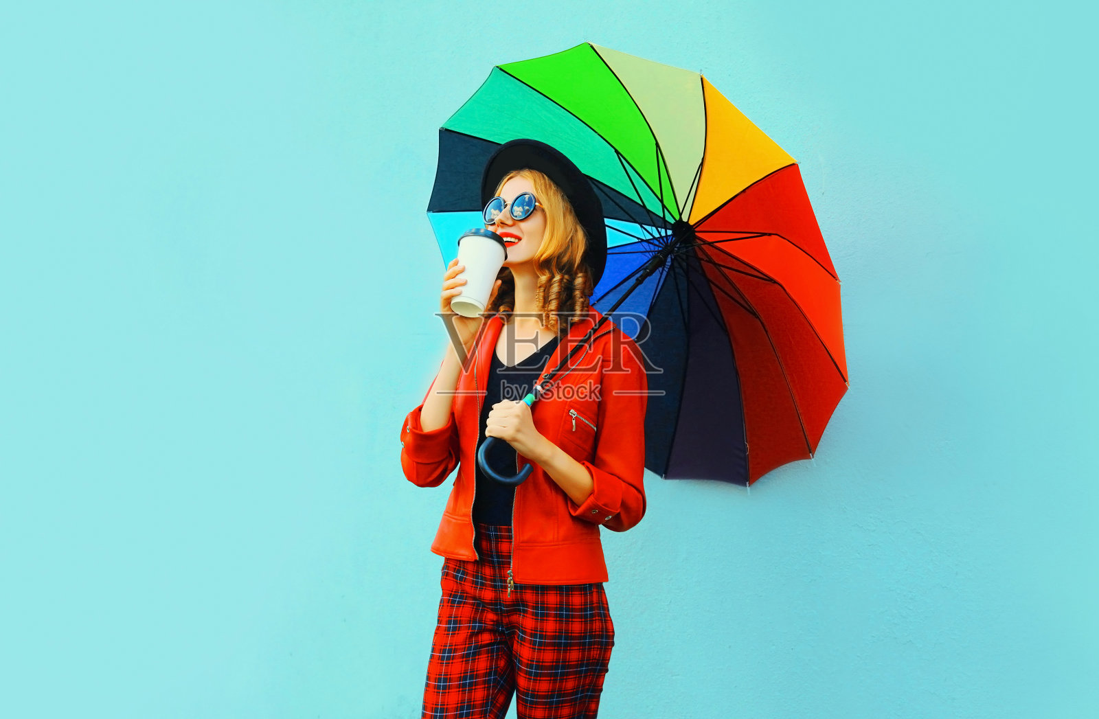 年轻女子喝着咖啡，拿着五颜六色的伞走在红色夹克，黑色的帽子在蓝色的墙壁背景照片摄影图片