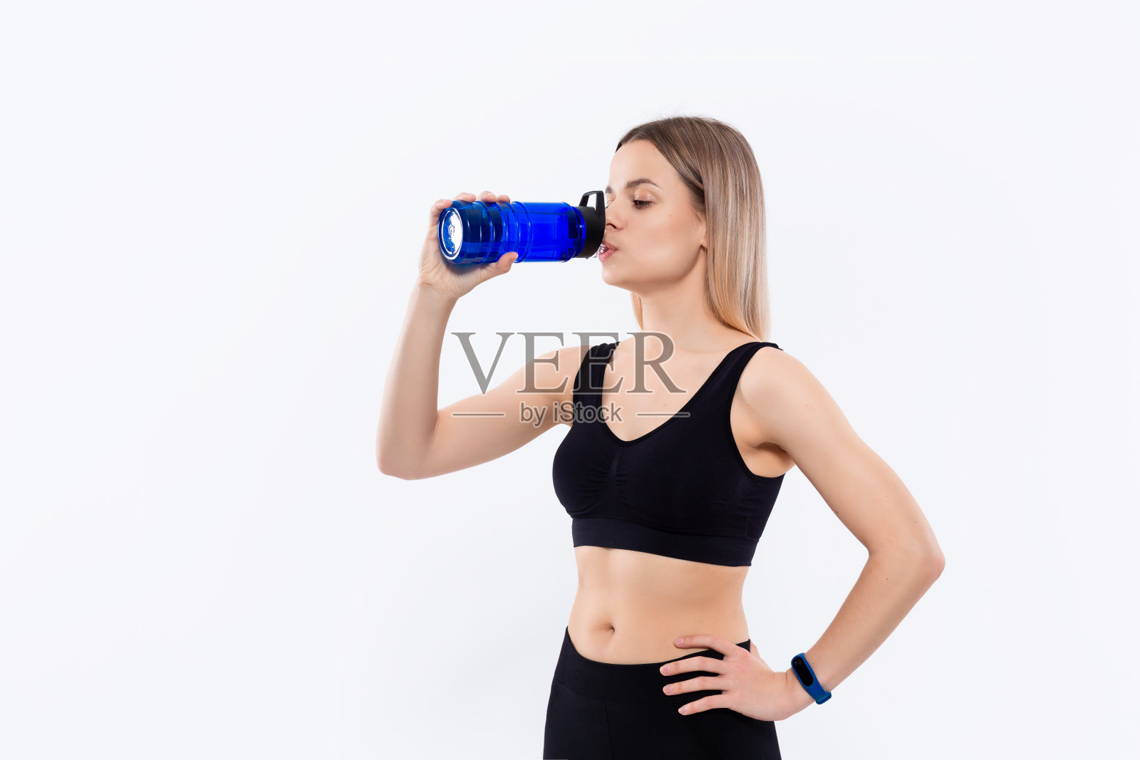 年轻的运动金发女人穿着黑色运动服与智能手表饮料后，站在白色的背景。照片摄影图片