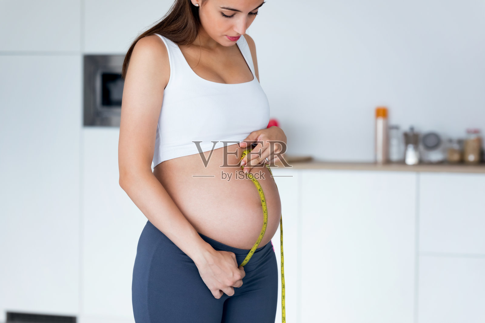 美丽的孕妇用卷尺测量自己的腹部来跟踪胎儿的发育。健康怀孕的概念。照片摄影图片