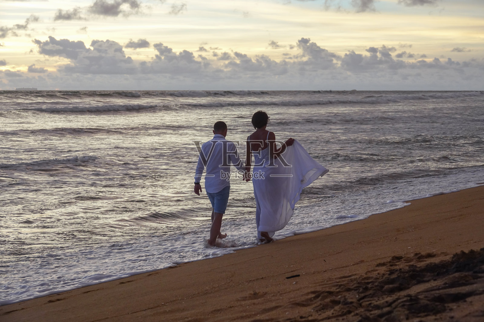 日落时分，一对相爱的年轻情侣手牵着手漫步在海滩上照片摄影图片