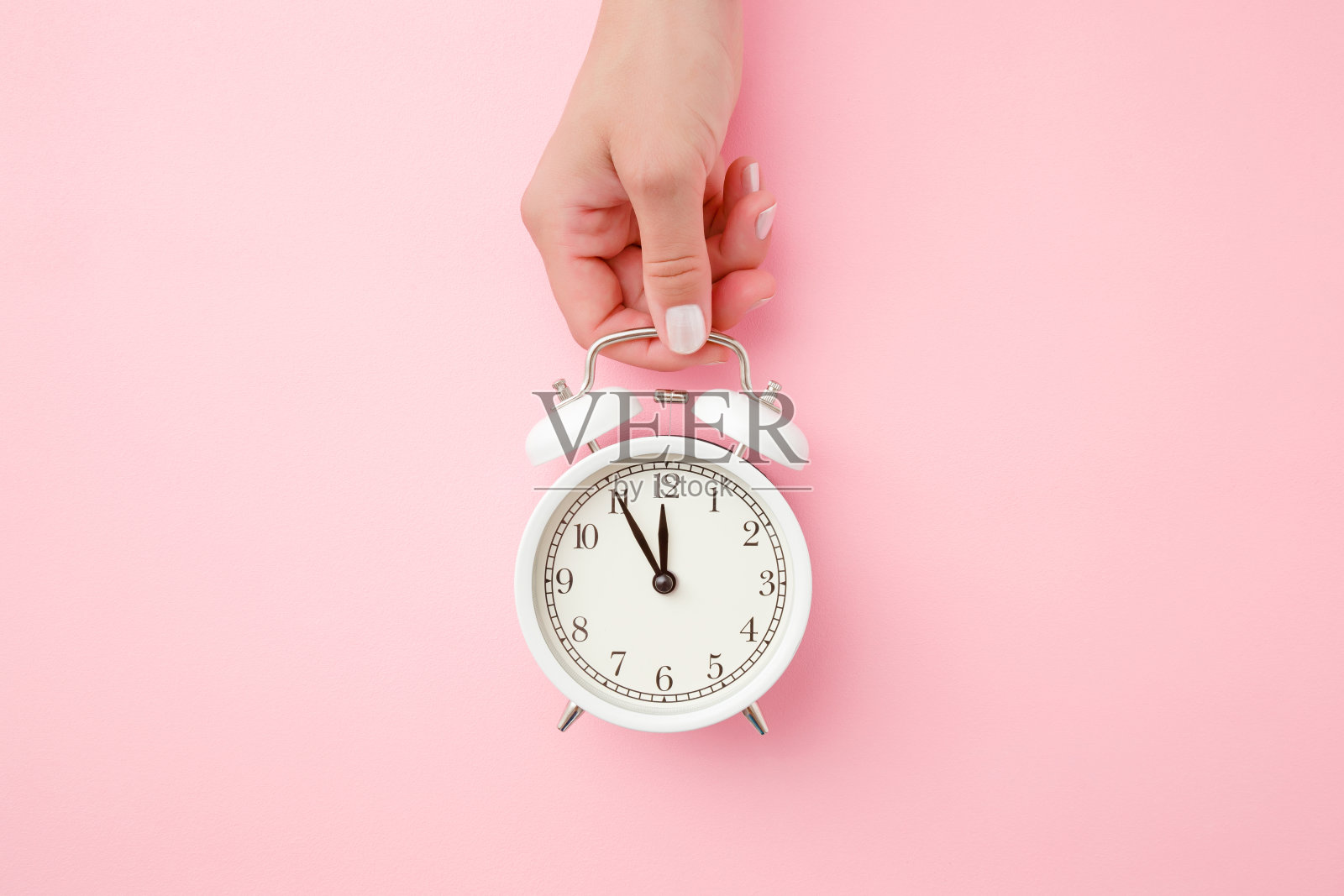 在淡粉色的背景下，一个女人手里拿着一个白色的闹钟。时间的概念。特写镜头。前视图。照片摄影图片