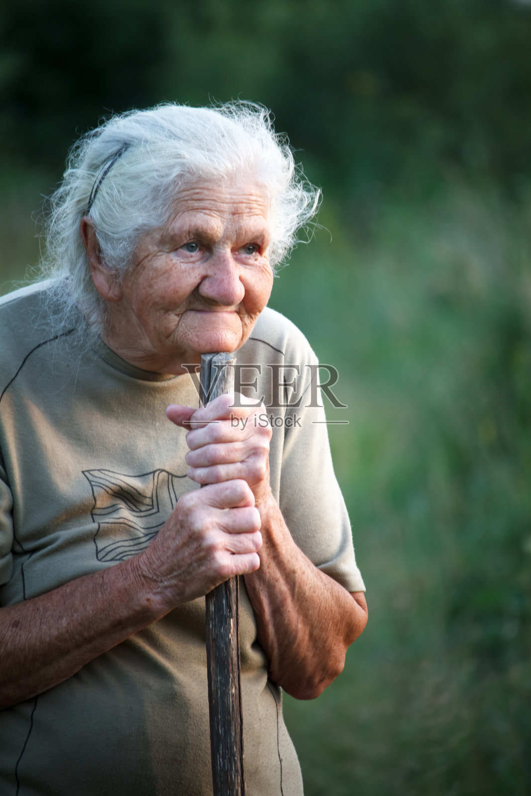 一个灰白头发的老妇人的特写，微笑着向上看，她的下巴搁在一根拐杖上，好像在拄着拐杖走路，脸上有深深的皱纹，选择性聚焦照片摄影图片