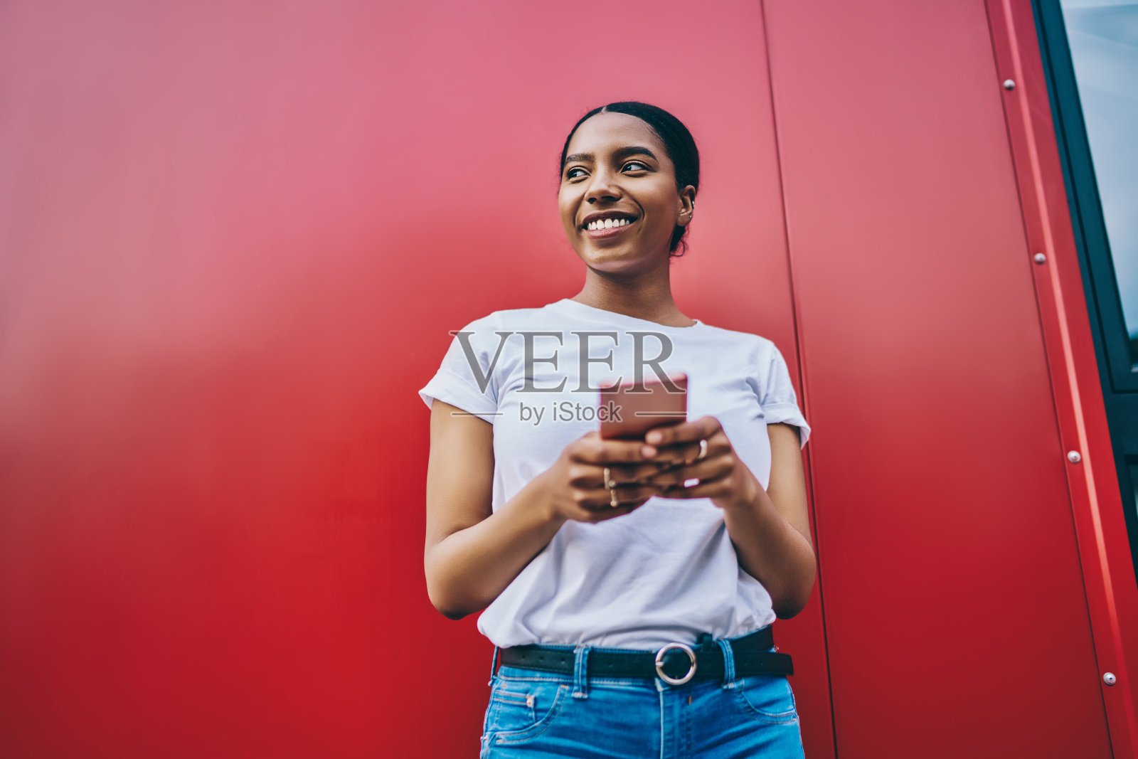 微笑的年轻非洲裔美国妇女在t恤拿着手机和看向远处，开朗的黑皮肤女博主用智能手机站在红色的墙背景。在线网络连接照片摄影图片