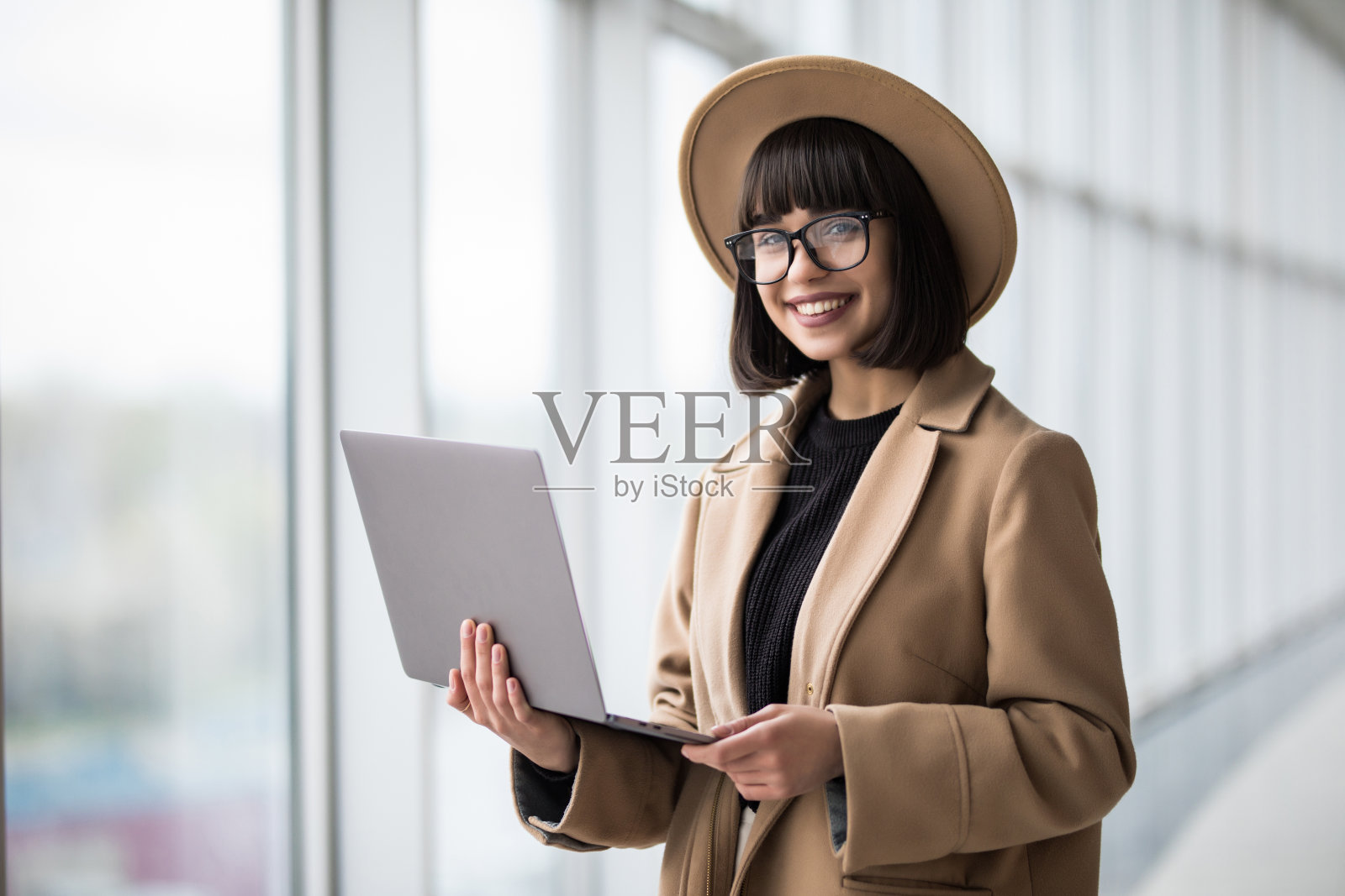年轻漂亮的女人穿着大衣和帽子在办公室靠窗使用笔记本电脑照片摄影图片