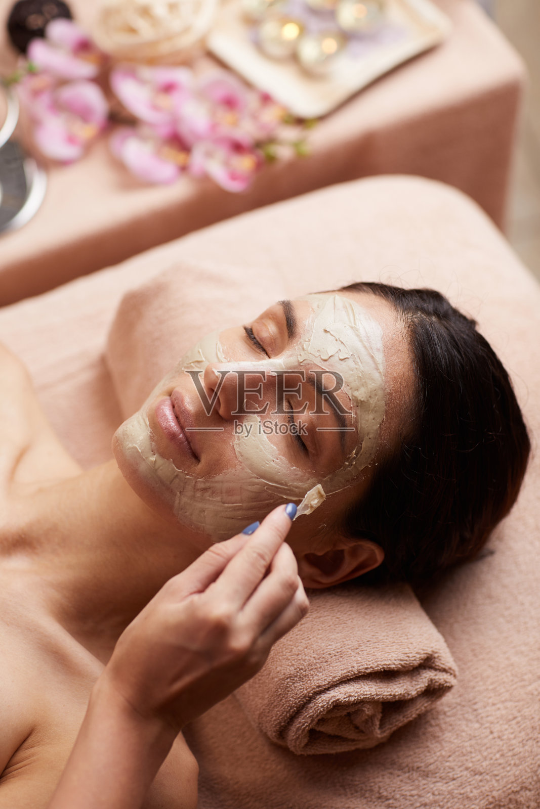 放松年轻的黑发女子躺在舒适的桌子上，在头下毛巾，而美容师敷面膜给她照片摄影图片