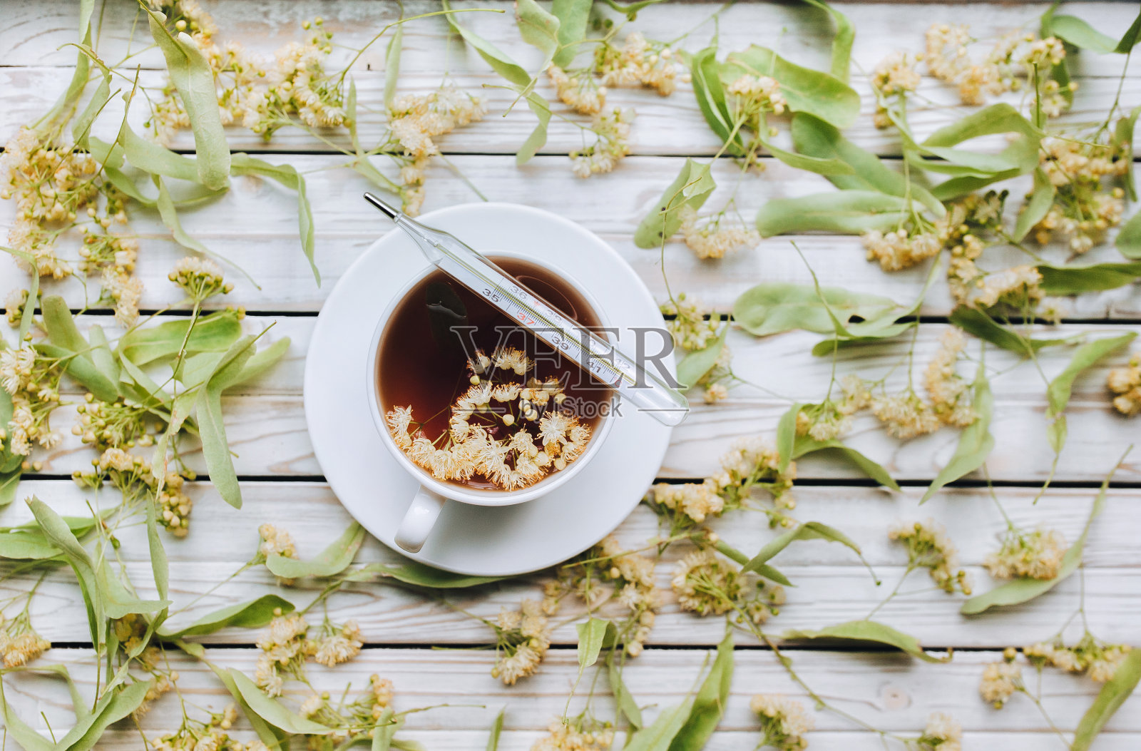 白色的杯子上放着一个水银玻璃温度计，上面放着一种由干菩提花制成的芳香治疗茶，放在灰色的木质背景上。草药的概念。照片摄影图片