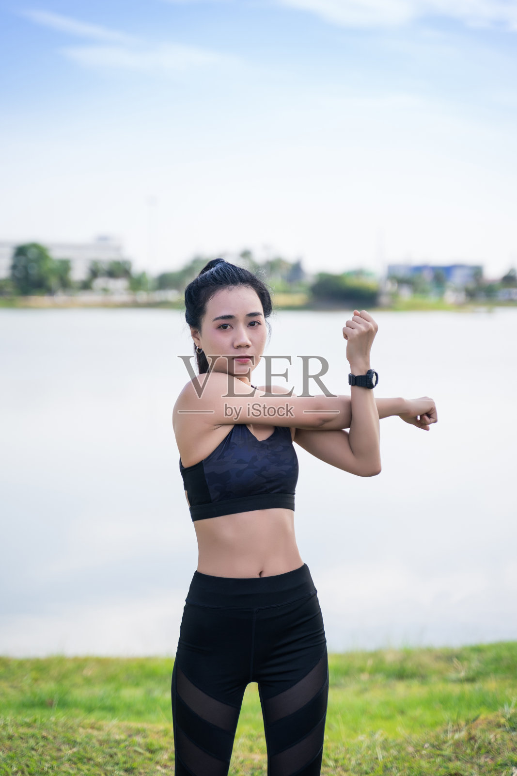 一个年轻的亚洲女人伸展前后健身运动在湖边的公园。一位年轻漂亮的女性穿着运动服在公园户外做伸展运动。正在户外热身的健康女性。照片摄影图片