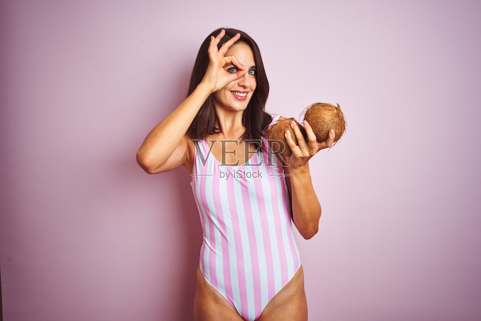 美丽的女人穿着粉红色条纹泳衣拿椰子在孤立的粉红色背景与快乐的脸微笑做ok手势与手在眼睛看通过手指照片摄影图片