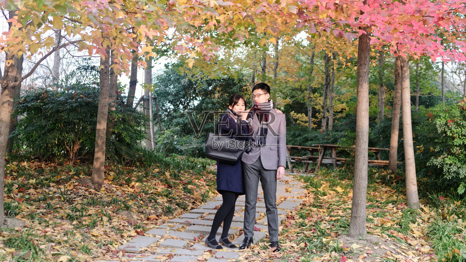 一个美丽的中国年轻女子拥抱她的男朋友的肩膀在秋天的森林公园，温暖的阳光照亮他们的脸，浪漫的时刻。照片摄影图片