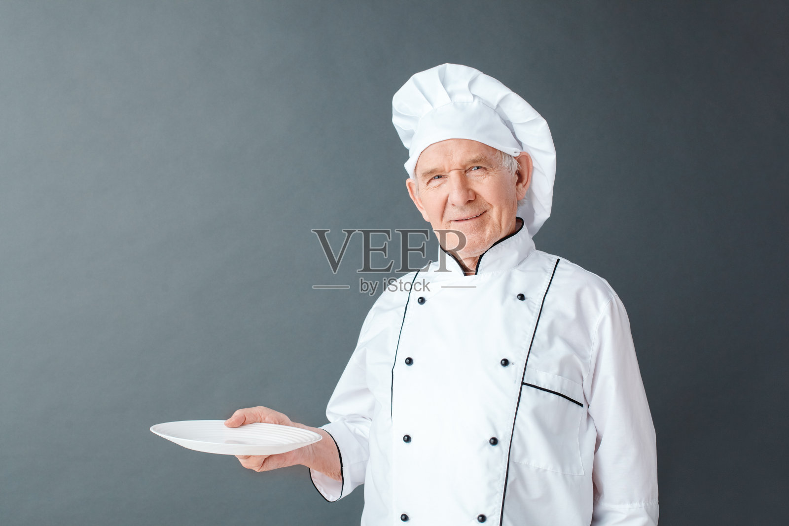 高级厨师长孤立地站在灰色的工作室里，用空盘子看着相机开心照片摄影图片