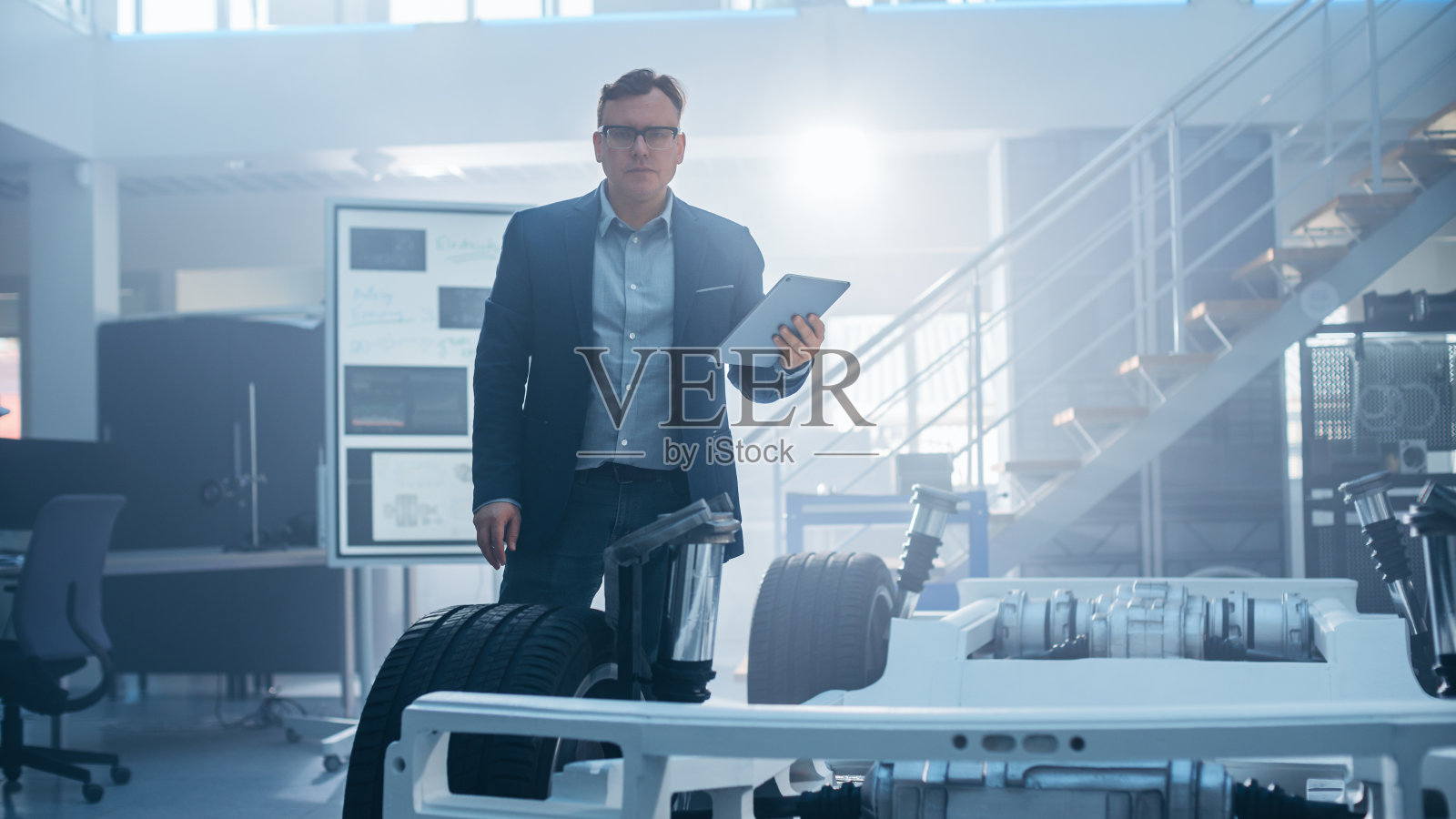 在高科技开发实验室里，一名戴眼镜的工程师在平板电脑上工作，旁边是一辆带轮子、电池和引擎的电动汽车底盘原型车。照片摄影图片