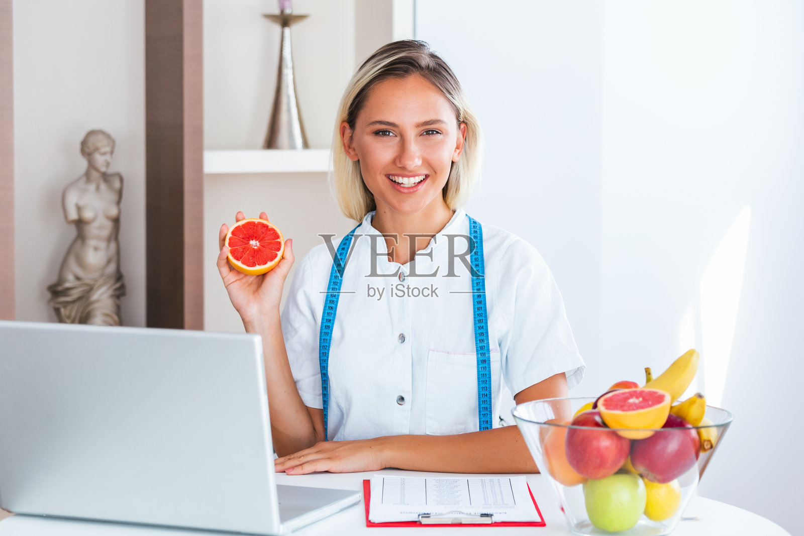 微笑的营养师在她的办公室，她展示了健康的蔬菜和水果，保健和饮食的概念。女营养学家，水果在办公桌前工作。照片摄影图片