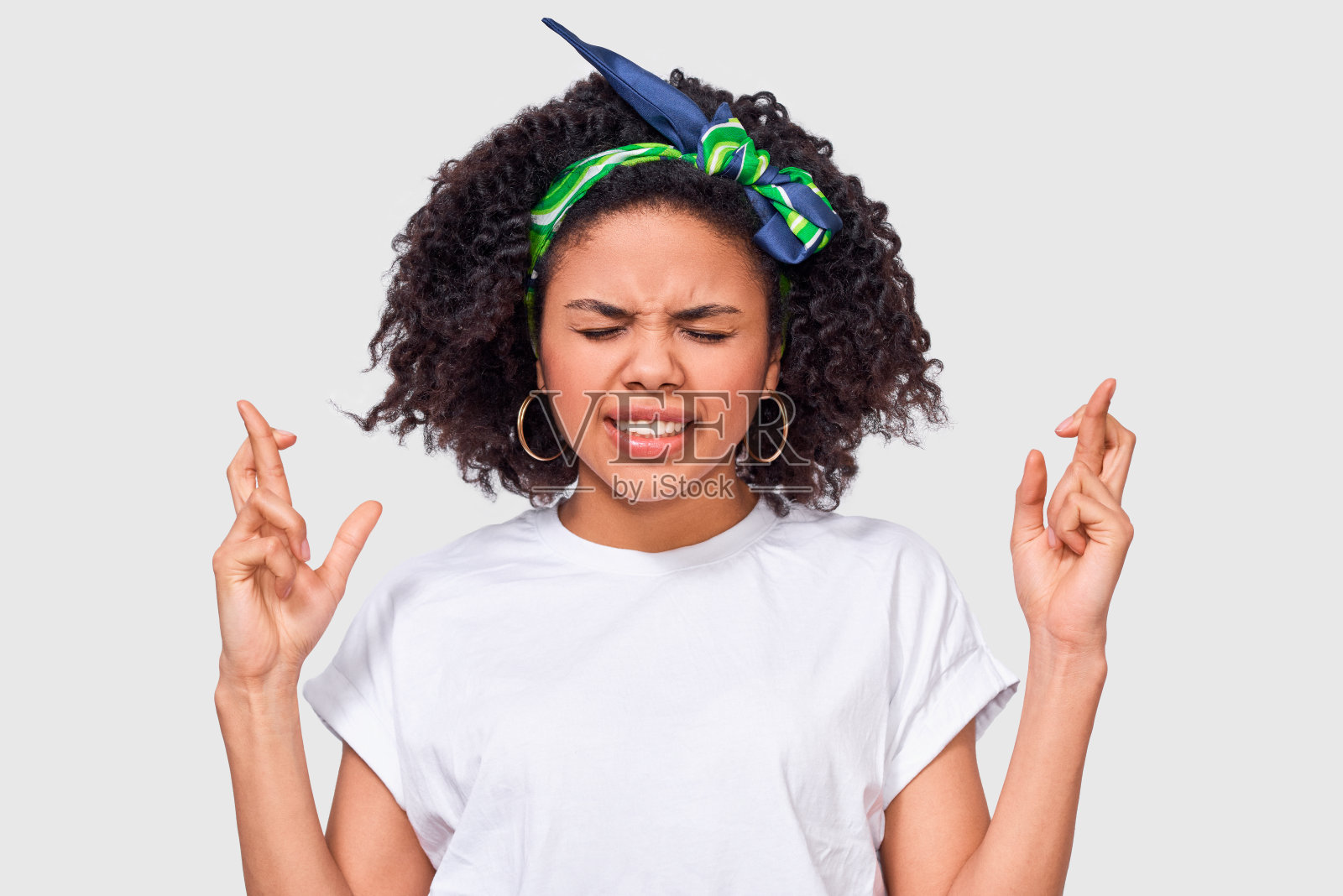 年轻的非裔美国妇女咬紧牙关，竖起手指，祈祷，穿着休闲的白色t恤。一厢情愿的女性交叉手指，在工作室里摆姿势。照片摄影图片