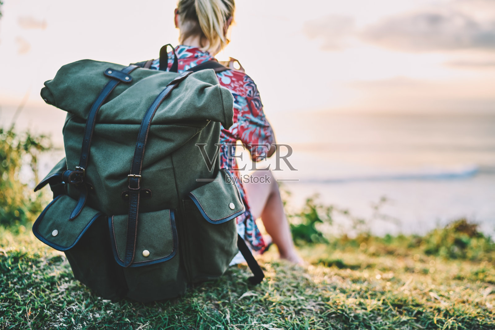 后视图的女人旅行者穿着休闲衣服放松在黄昏时间坐在大背包旁边的海洋。裁剪图像的女性游客再创造，而花时间在海附近照片摄影图片