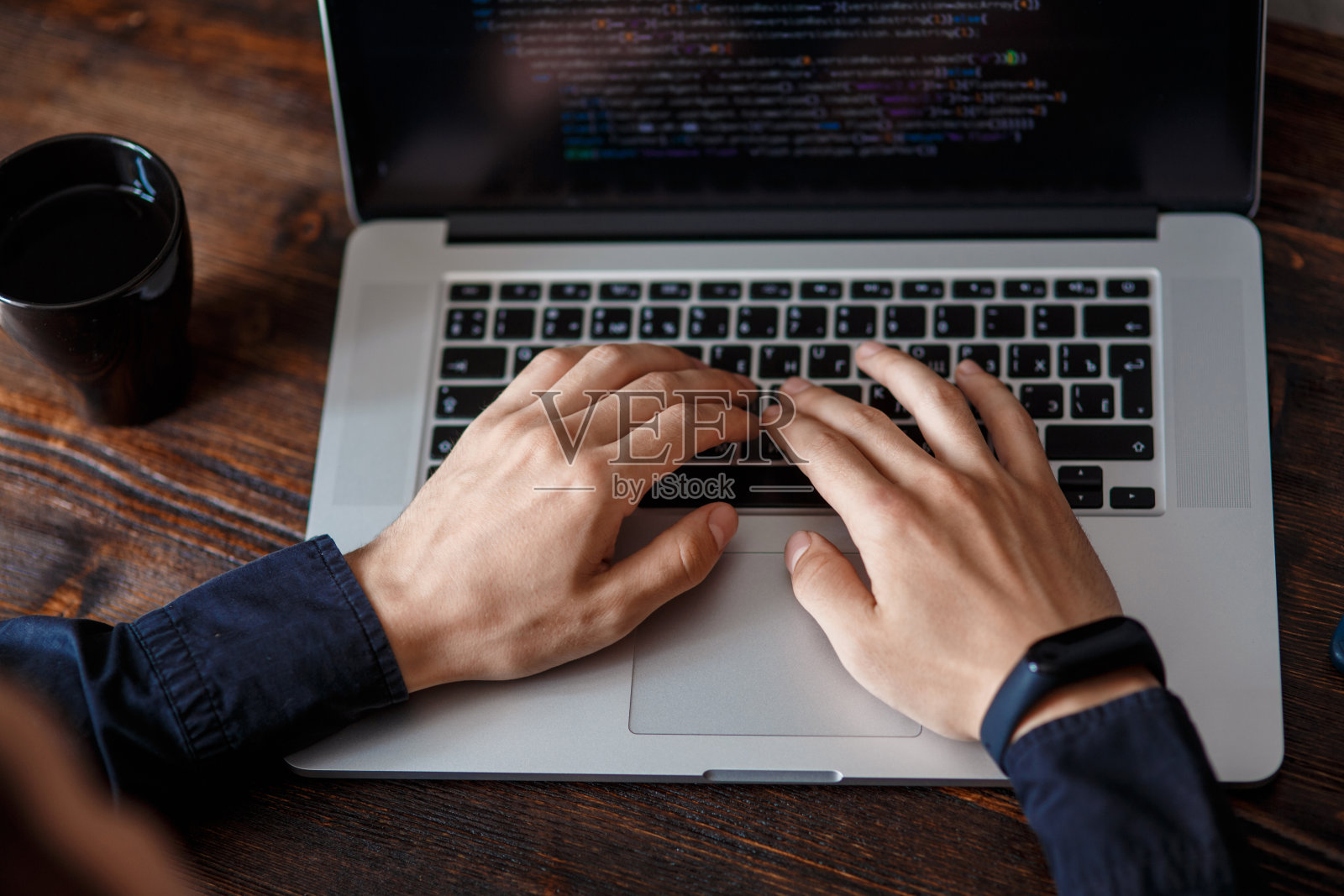 一名身穿黑衬衫的男程序员正在一台笔记本电脑前工作，电脑上放着程序代码和开发人员的双手。照片摄影图片