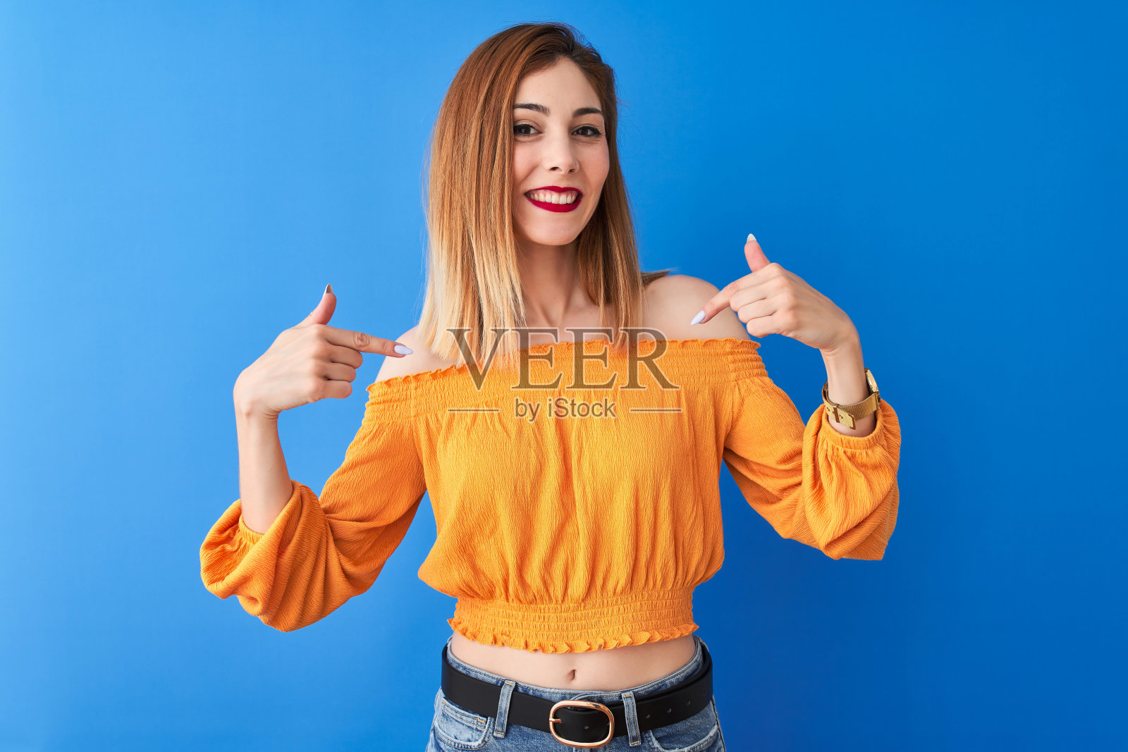 美丽的红发女人穿着橙色休闲t恤站在孤立的蓝色背景看起来自信的微笑在脸上，用手指指着自己骄傲和幸福。照片摄影图片