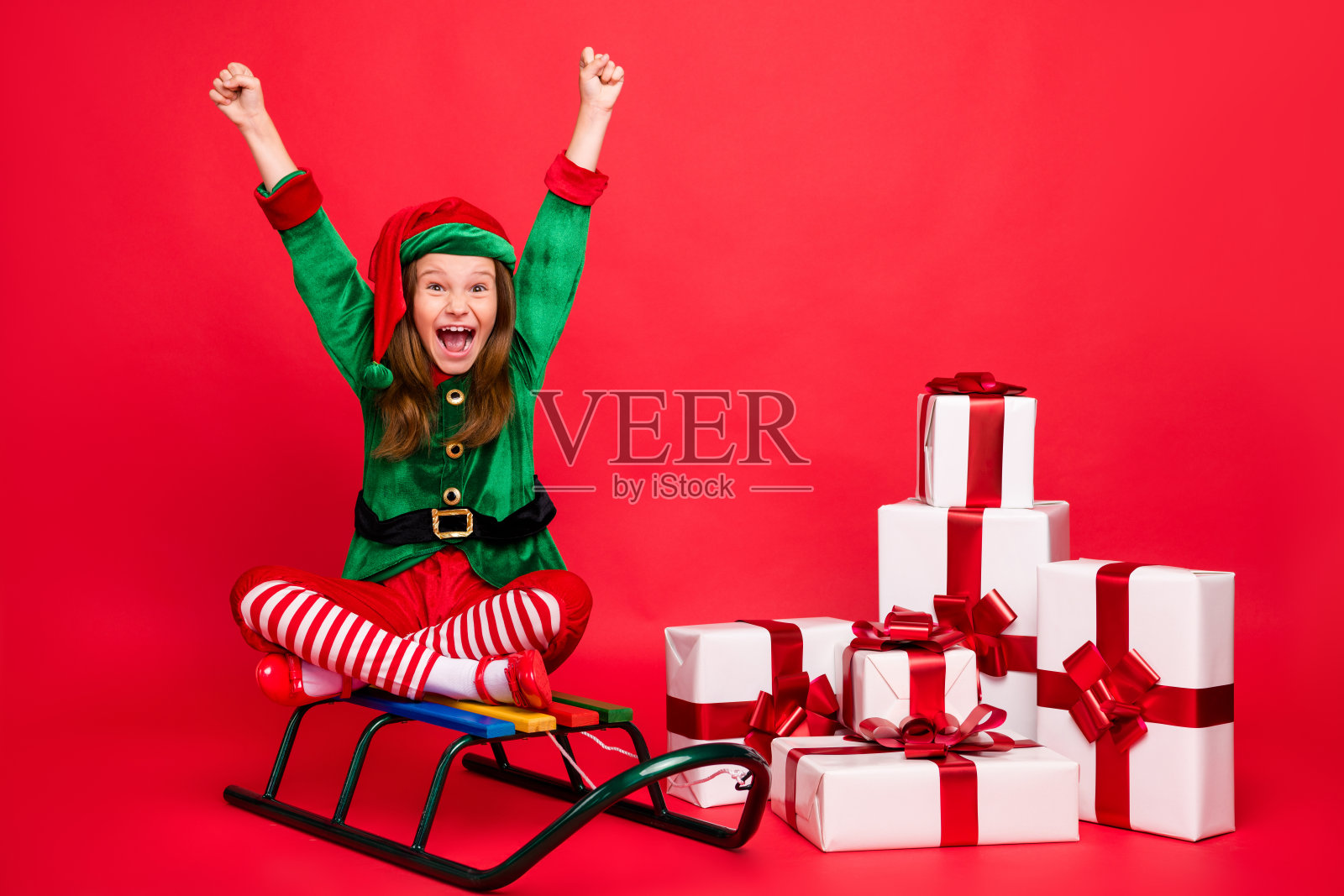 她的肖像，她漂亮迷人，快乐，喜出风头的十多岁的精灵圣诞老人，看坐在雪橇上，莲花的姿势姿势，举起双手，高兴孤立在明亮生动的照耀红色的背景照片摄影图片
