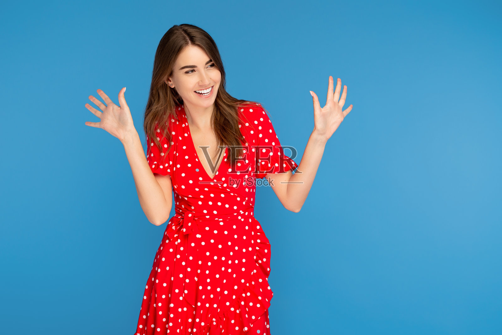美丽的年轻女子与明亮的微笑在红色休闲连衣裙向上看与兴奋的脸与手在蓝色的背景。照片摄影图片