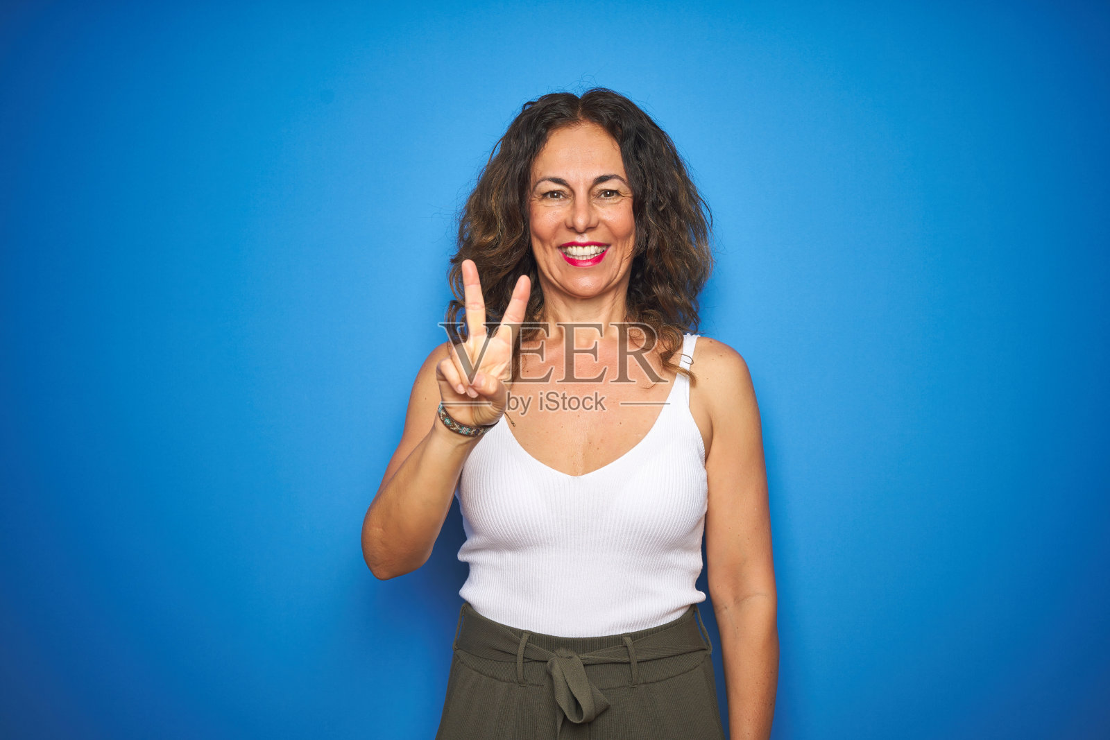 一位卷发的中年妇女站在蓝色孤立的背景下，用第二根手指向上指，同时微笑着，自信而快乐。照片摄影图片