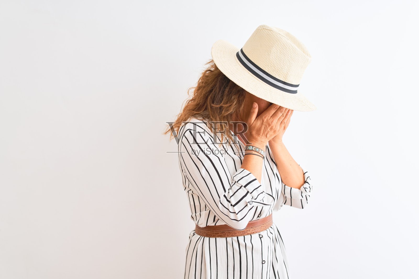 身穿条纹连衣裙、戴帽子的中年女商人站在白色背景前，用手捂住脸哭泣，神情悲伤，表达沮丧照片摄影图片