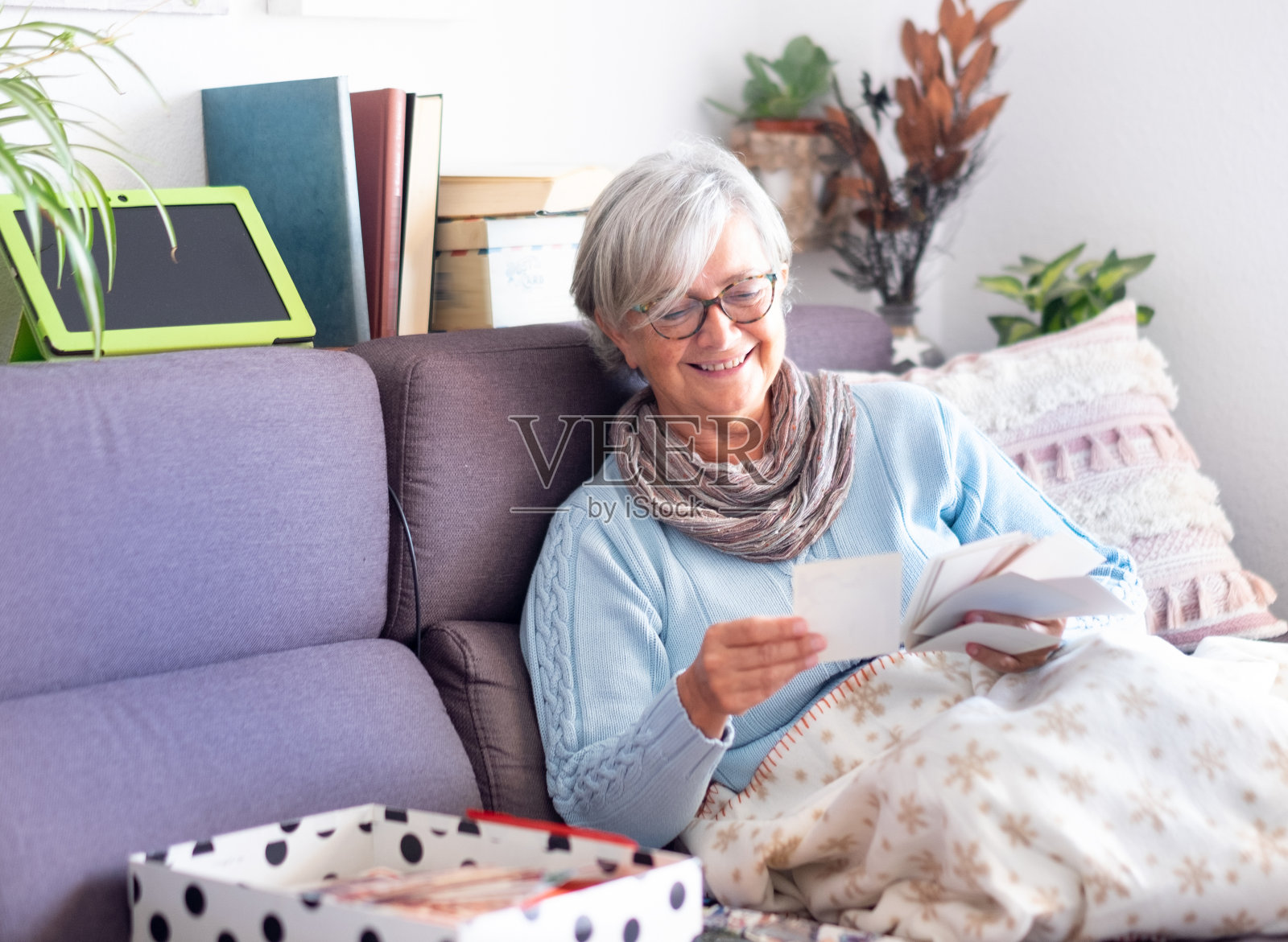 正面的观点，一个有吸引力的资深女士看老照片的一生微笑。坐在沙发上，用手机和平板电脑。白色的墙壁和书的背景照片摄影图片