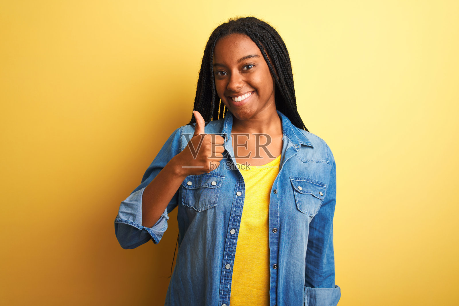 年轻的非洲裔美国妇女穿着牛仔衬衫站在孤立的黄色背景做快乐的拇指手势与手。赞许的表情看着镜头，表示成功。照片摄影图片