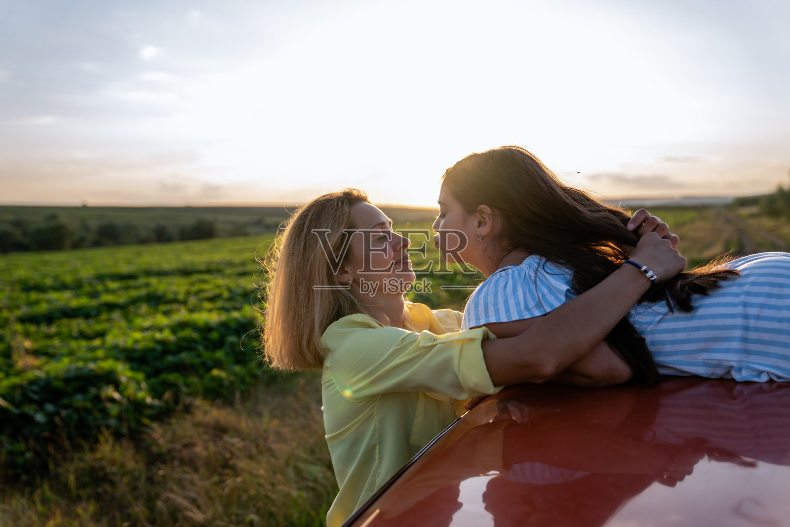 穿着黄色衬衫的年轻妈妈拥抱着她，看着她十几岁的女儿，她穿着蓝色条纹的裙子躺在一辆红色汽车的车顶上，看着她的妈妈，夕阳在背景中照片摄影图片