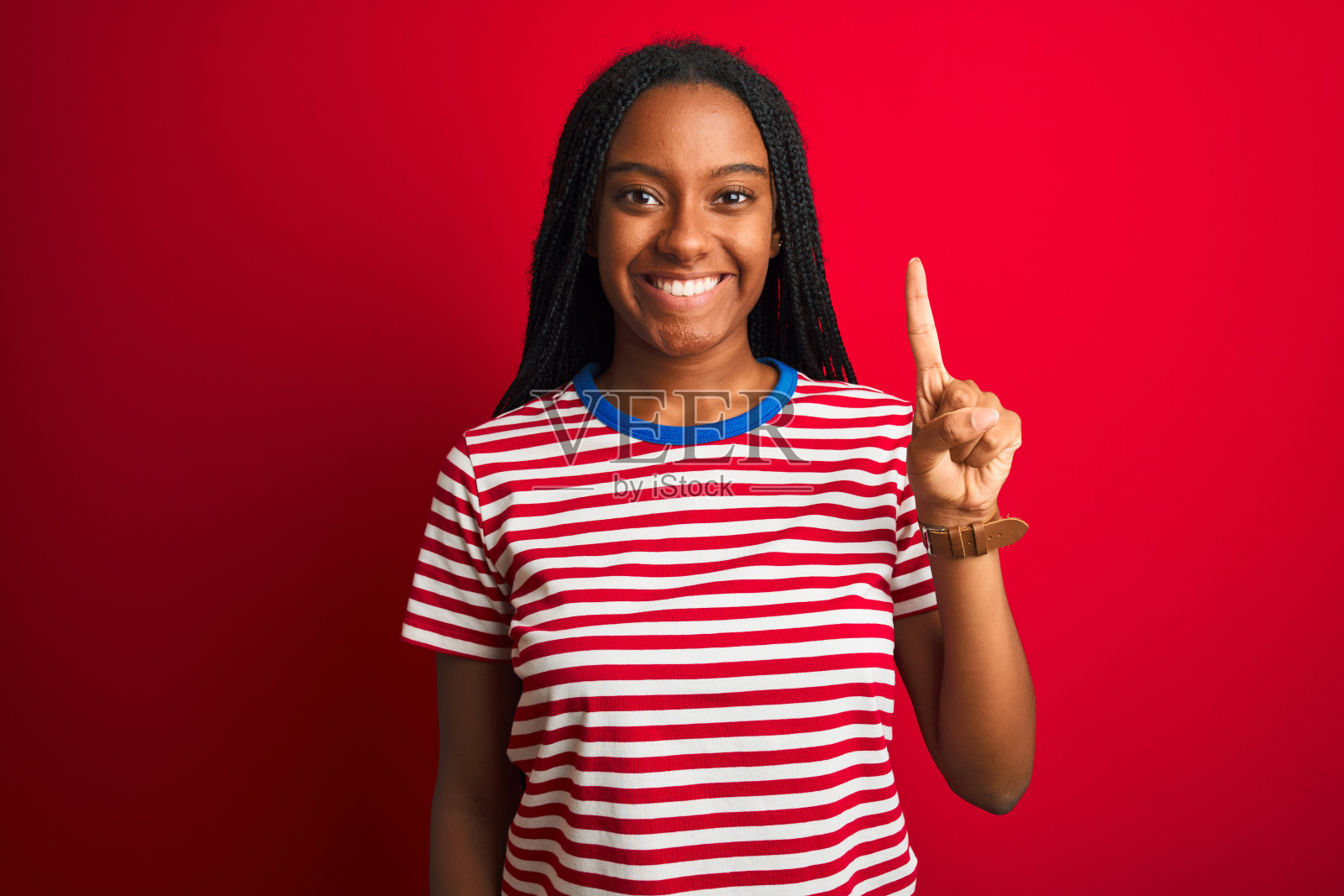年轻的非洲裔美国妇女穿着条纹t恤站在孤立的红色背景显示和指向第一个手指，同时自信和快乐的微笑。照片摄影图片