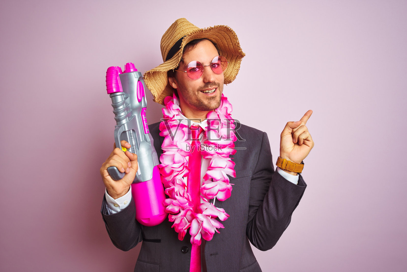 年轻的商人戴着水枪夏威夷花环帽眼镜在孤立的粉红色背景非常高兴地用手和手指指向一边照片摄影图片