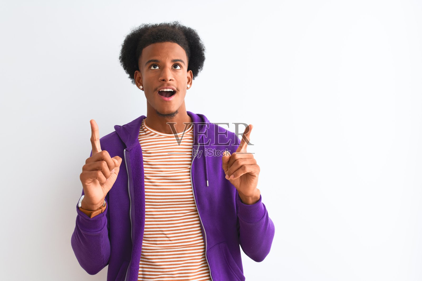 一名身穿紫色运动衫的非裔美国人站在孤立的白色背景下惊讶地抬头用手指指着并举起手臂。照片摄影图片