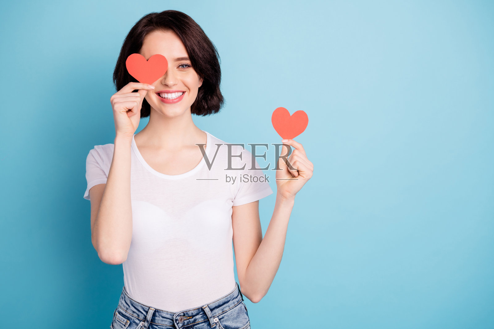 她的肖像，她漂亮的，迷人的，可爱的，快乐的，快乐的女孩手里拿着两个心符号卡，闭上眼睛，在明亮生动的闪耀，充满活力的蓝色绿松石色的背景上，有乐趣的孤立照片摄影图片