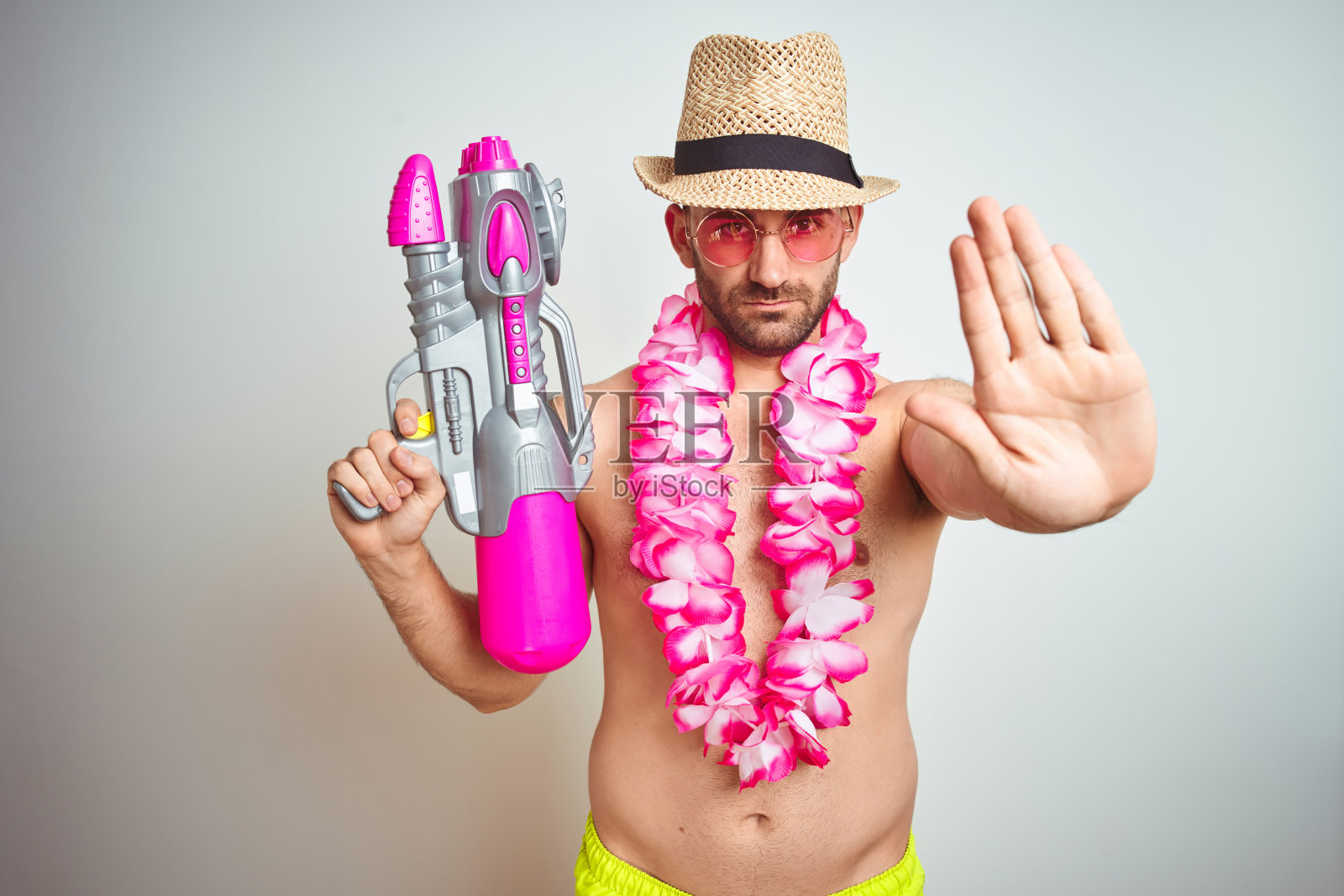 年轻男子戴着夏天的帽子和夏威夷花环拿着水枪在孤立的背景与开放的手做停止标志与严肃和自信的表情，防御手势照片摄影图片