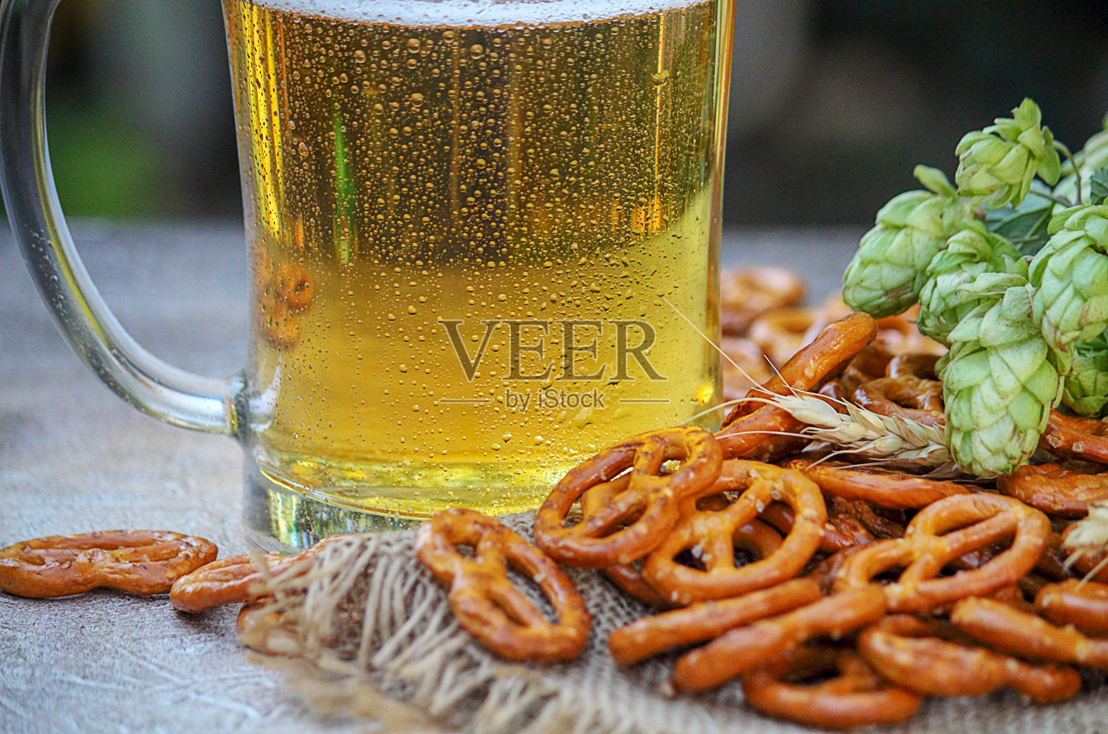 玻璃杯里的啤酒。一杯啤酒上的水滴。新鲜的绿啤酒花，小麦穗和椒盐卷饼摆在桌上。酿造和啤酒小吃的配料。照片摄影图片