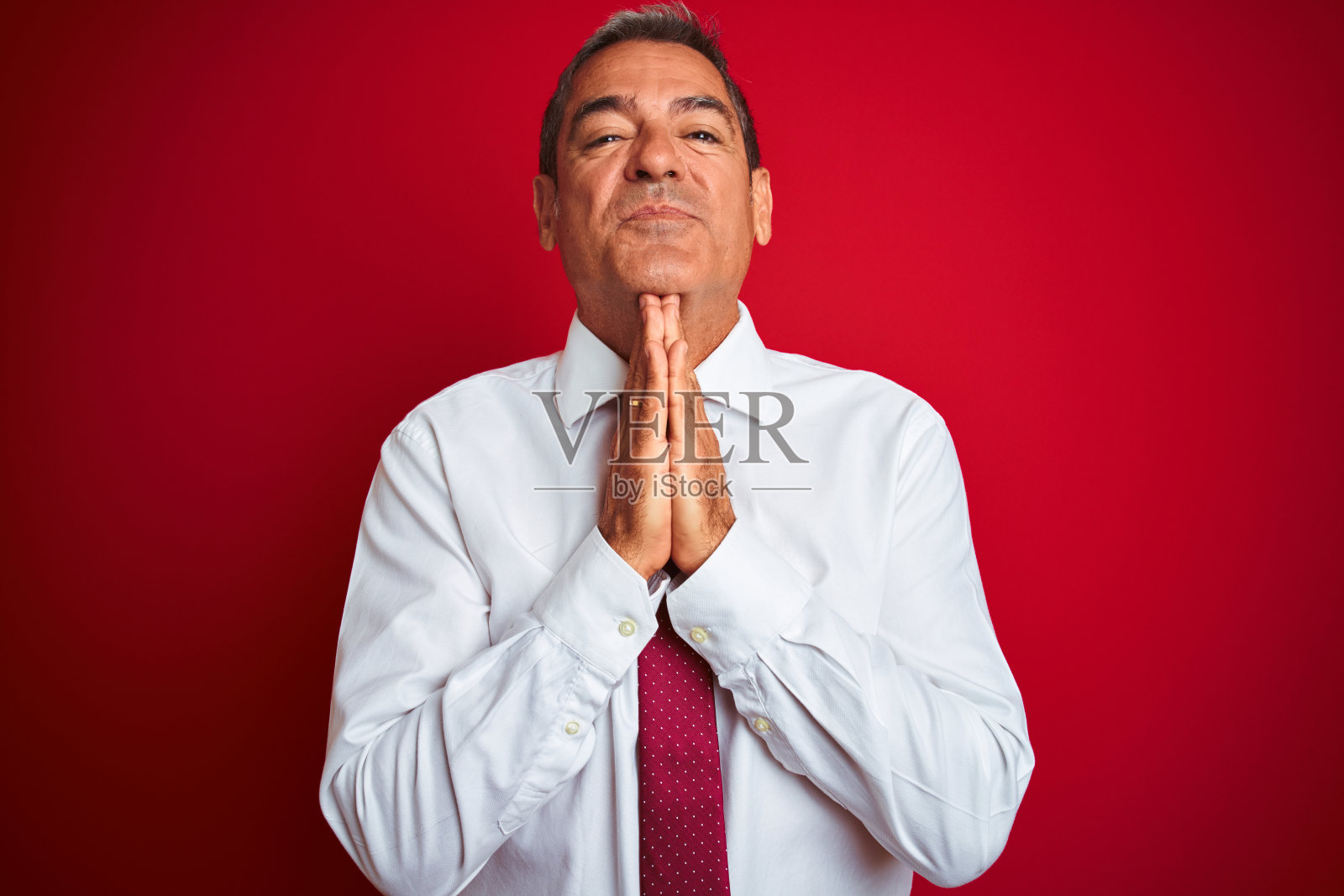 英俊的中年商人站在孤立的红色背景与手一起祈祷，请求原谅自信的微笑。照片摄影图片