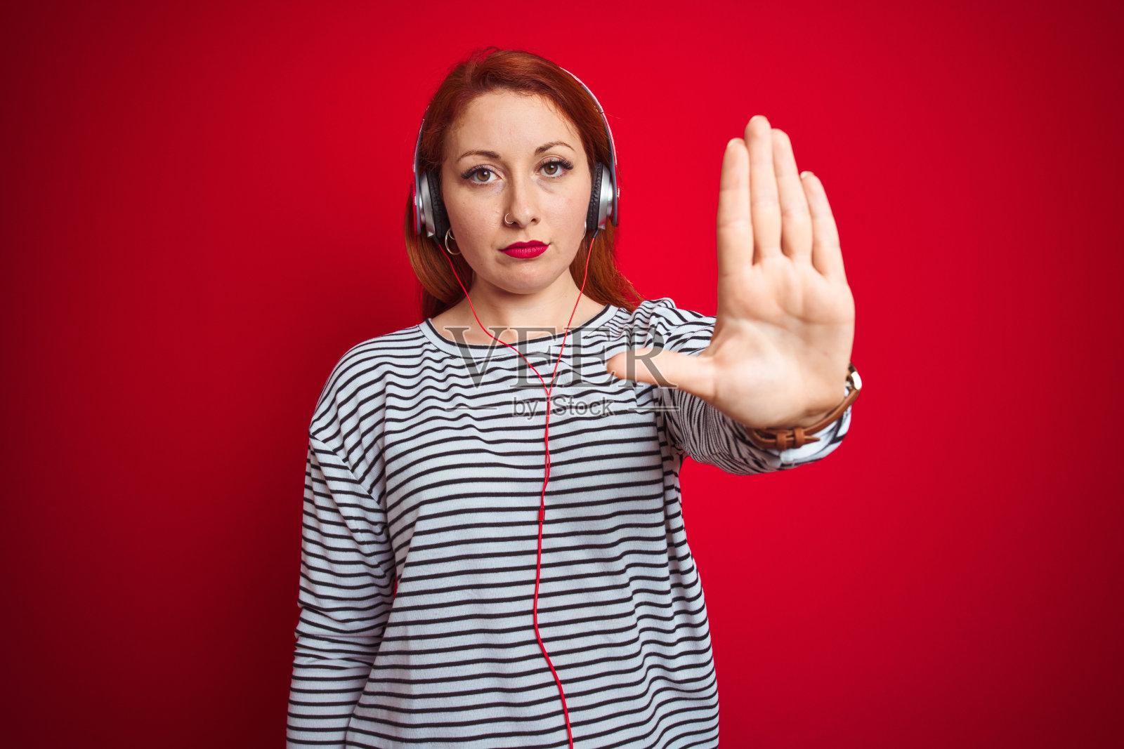 年轻的红发女子听音乐使用耳机在红色孤立的背景与开放的手做停止标志与严肃和自信的表情，防御手势照片摄影图片
