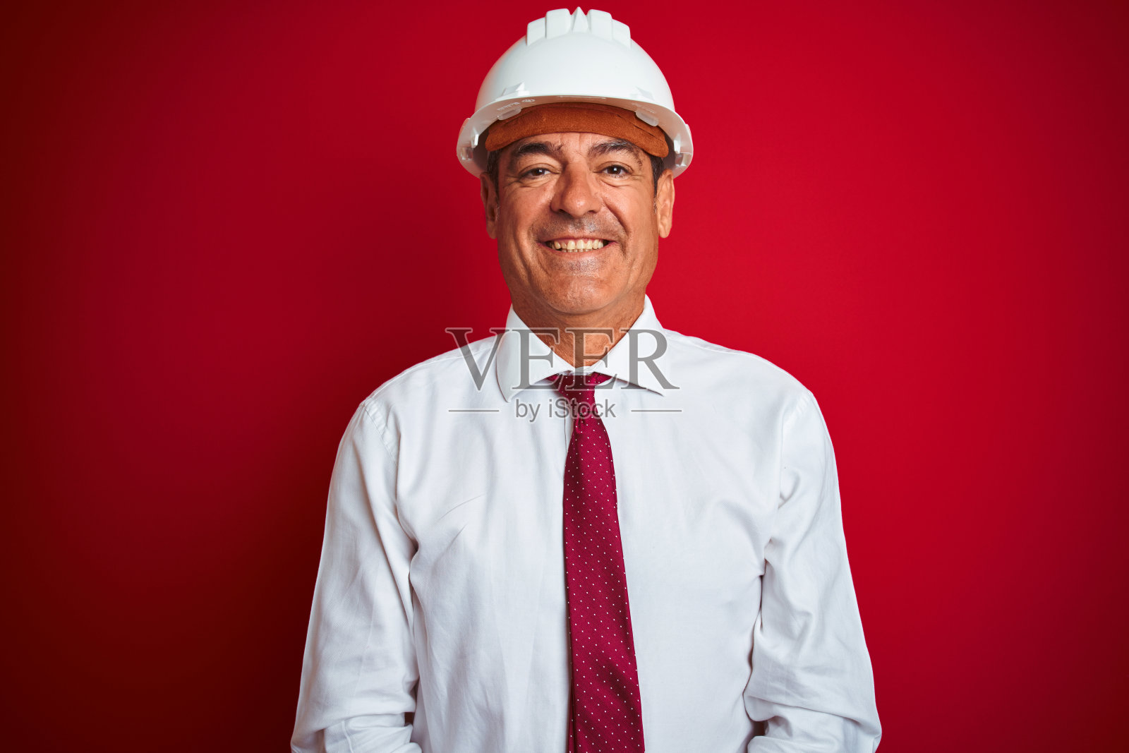 英俊的中年工程师男人戴着安全帽在孤立的红色背景与快乐和凉爽的微笑在脸上。幸运的人。照片摄影图片