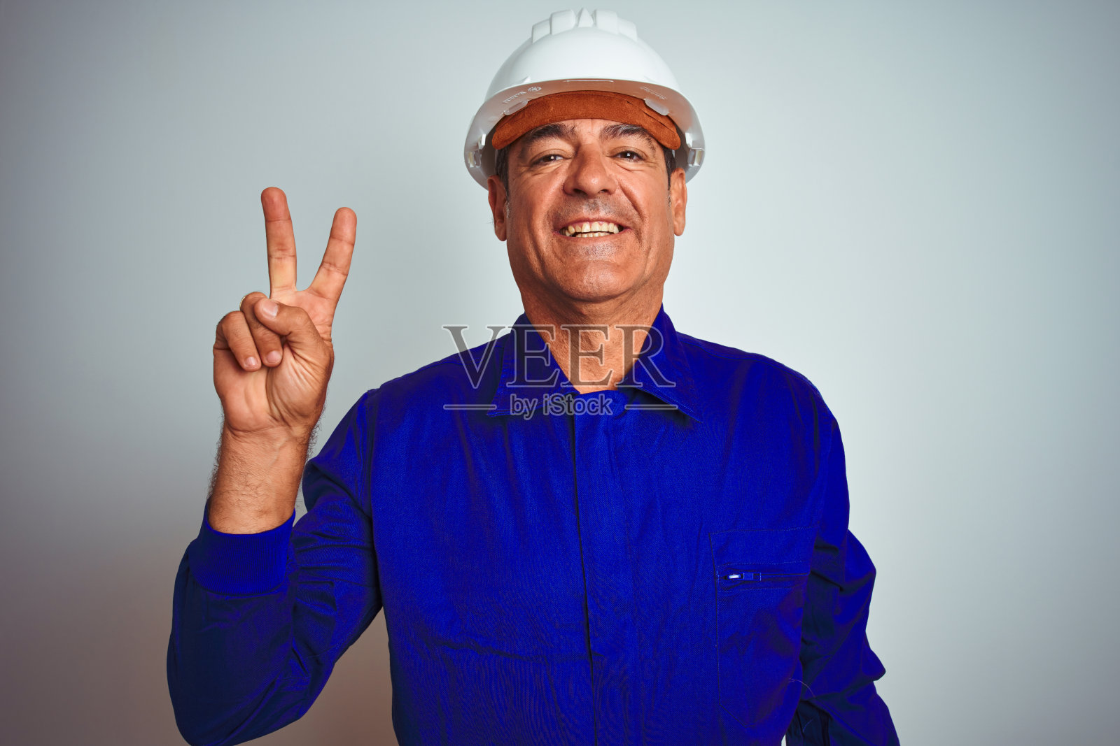 英俊的中年工人穿着制服和头盔在孤立的白色背景显示和指向2号手指，同时自信和快乐的微笑。照片摄影图片