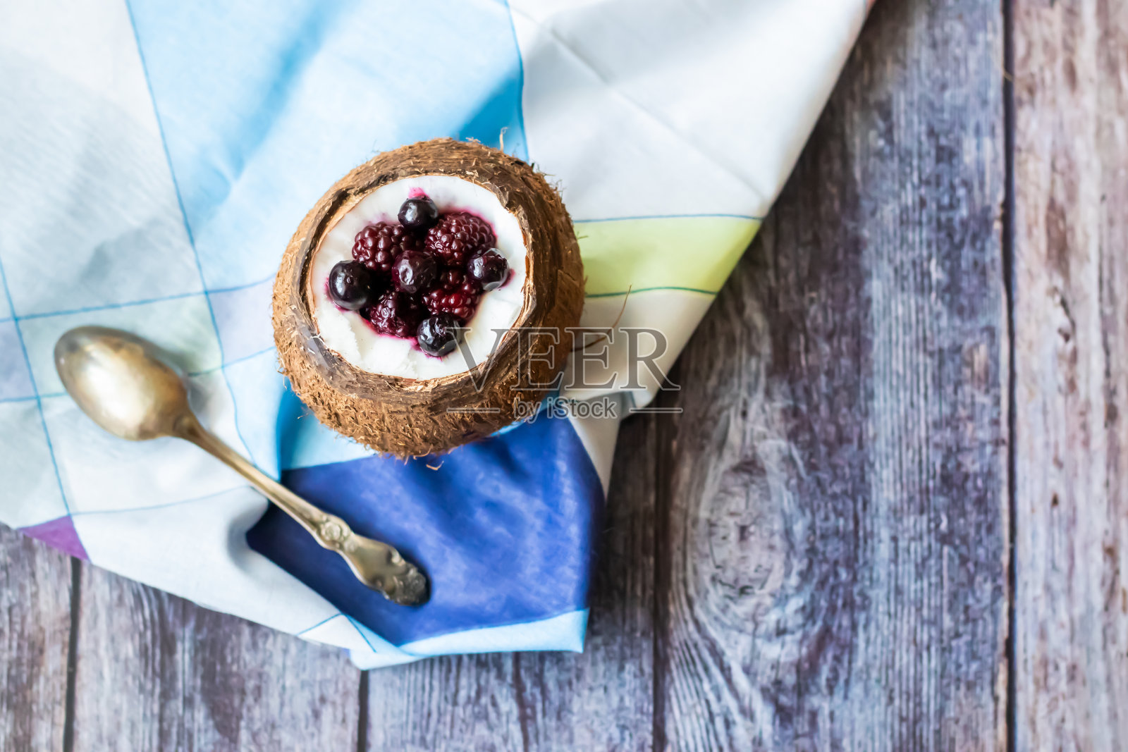 莓果甜点和酸奶在一杯椰子在木制的背景。黑莓，覆盆子，蓝莓，黑加仑子。蓝色的餐巾照片摄影图片
