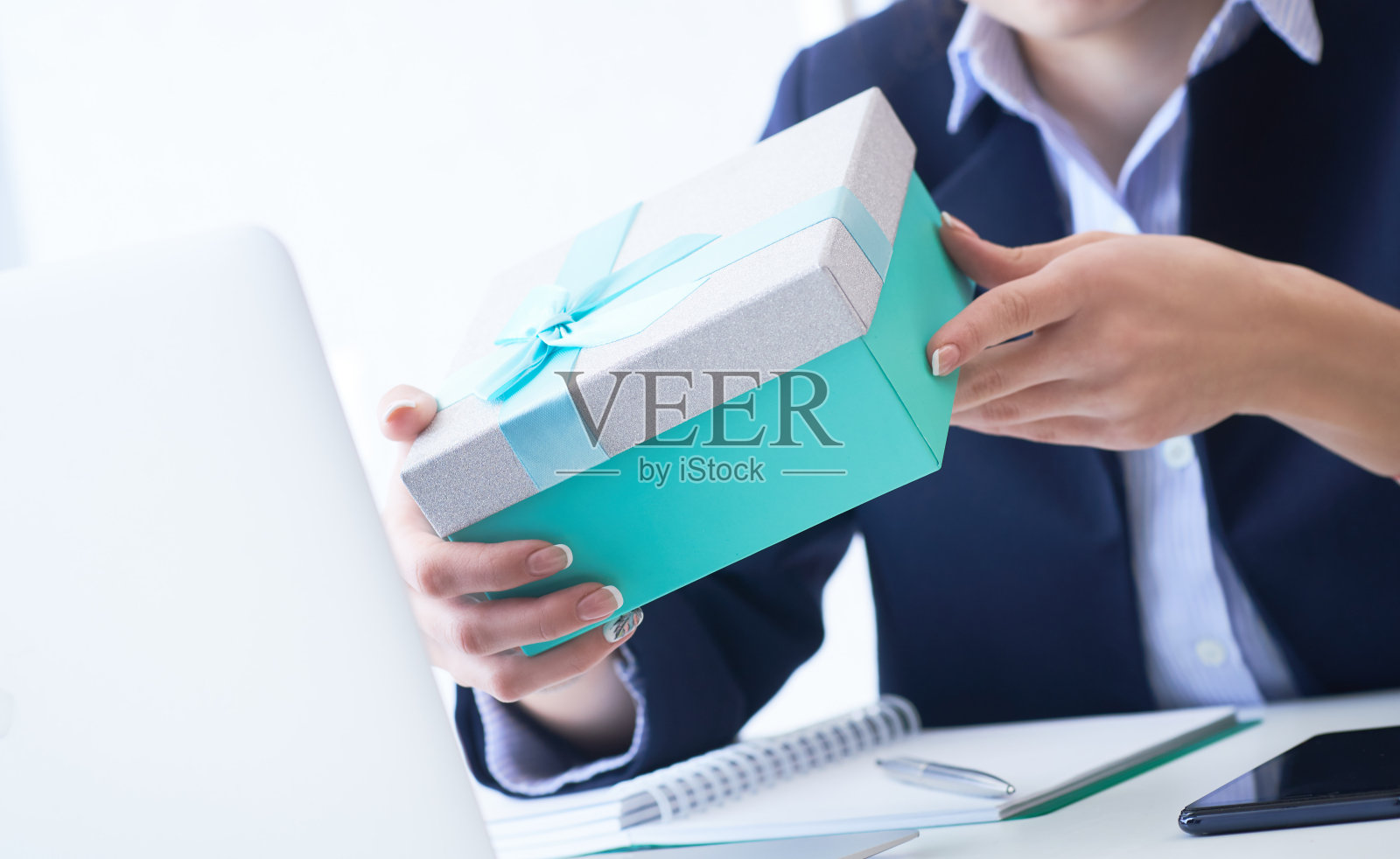 圣诞节,任何节日。一位办公室职员收到了同事送的礼物。特写女人的手拿着蓝色的礼盒在办公室背景。照片摄影图片