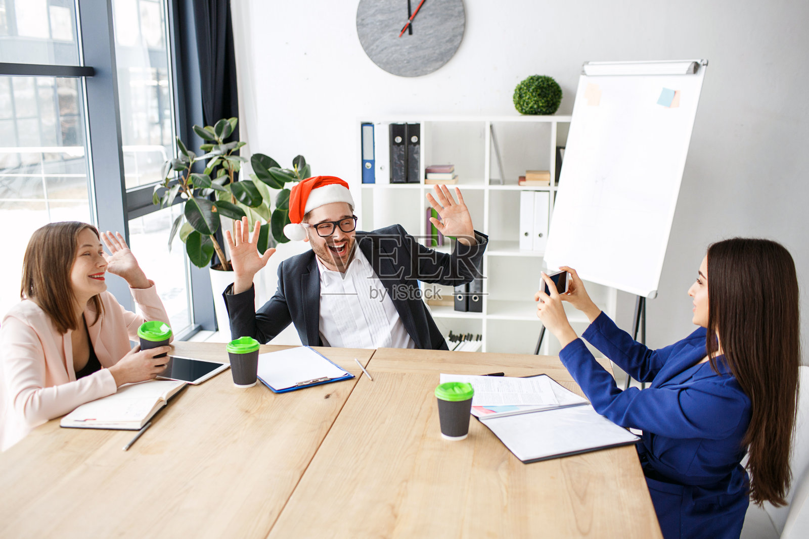 工人在办公室。庆祝新年或圣诞节。人们坐在一起吃饭。黑发女人在拍一个戴红帽子的男人。在工作中获得乐趣。照片摄影图片