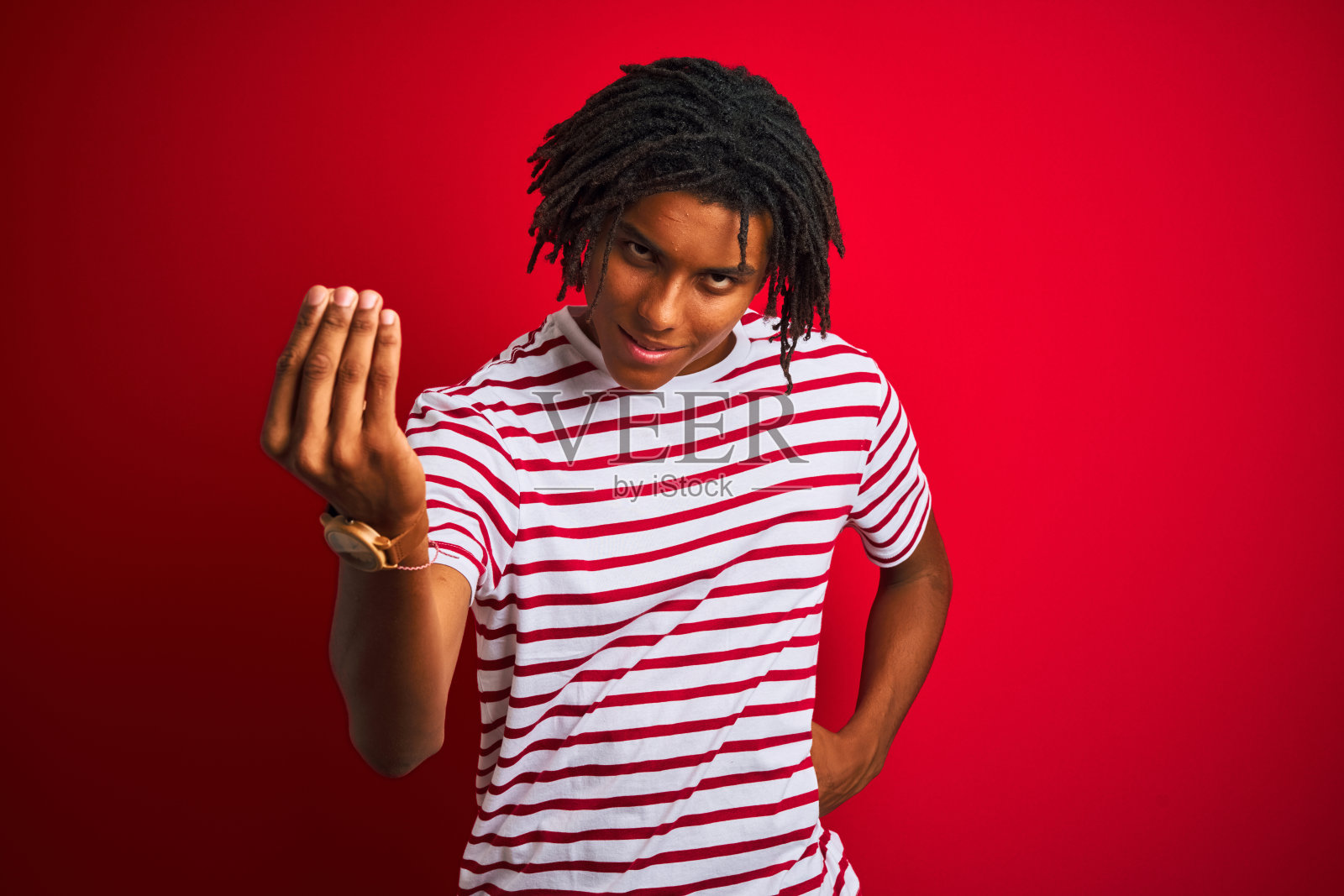 年轻的黑人，梳着雷鬼辫，穿着条纹t恤站在孤立的红色背景上，用手和手指做着意大利手势，充满自信的表情照片摄影图片