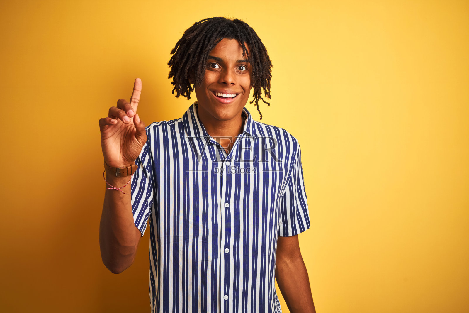 非洲人与雷鬼发穿着休闲条纹t恤孤立的黄色背景指向手指向上的成功的想法。退出和快乐。第一。照片摄影图片
