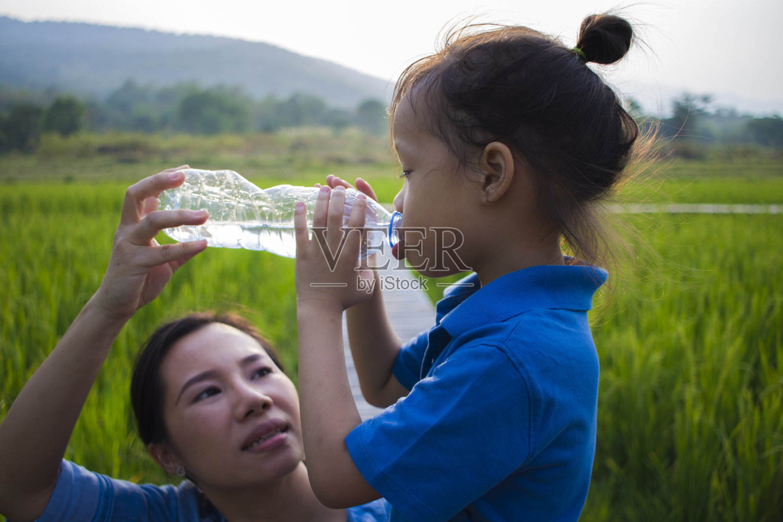 母亲帮助她的孩子们在稻田里喝瓶装水。长头发的男孩。照片摄影图片