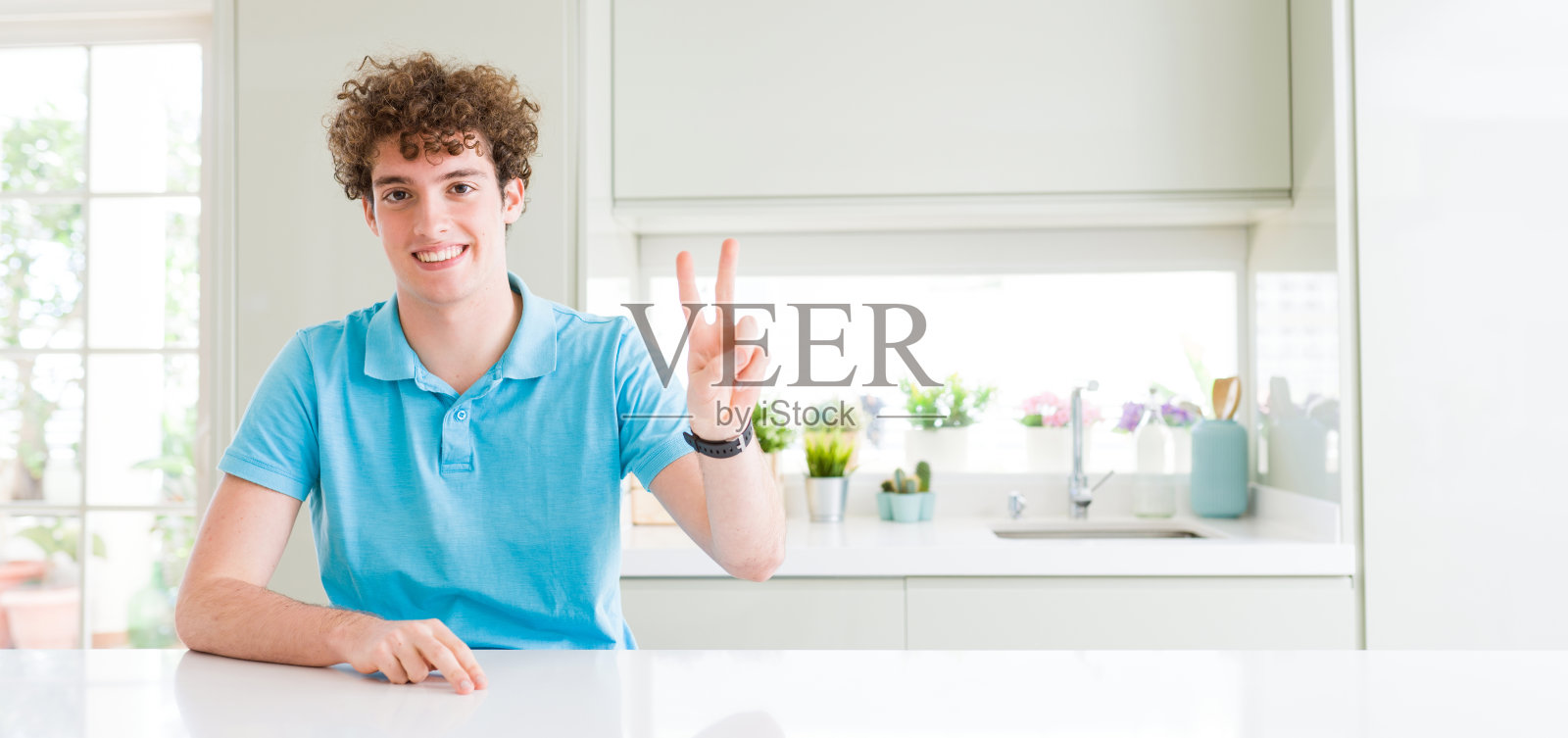 宽镜头的年轻英俊的男子在家里微笑看着镜头显示手指做胜利的手势。二号人物。照片摄影图片