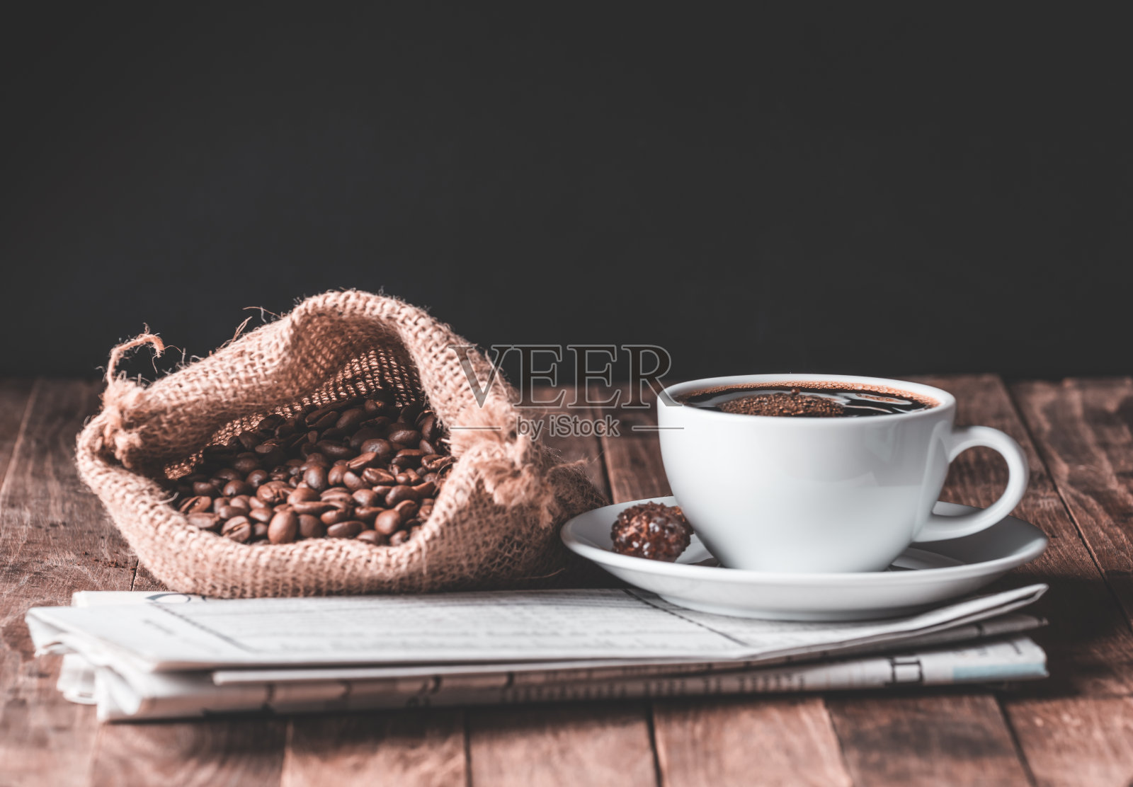 深色背景的木桌上摆放着咖啡杯、肉桂棒、烘咖啡豆和报纸照片摄影图片