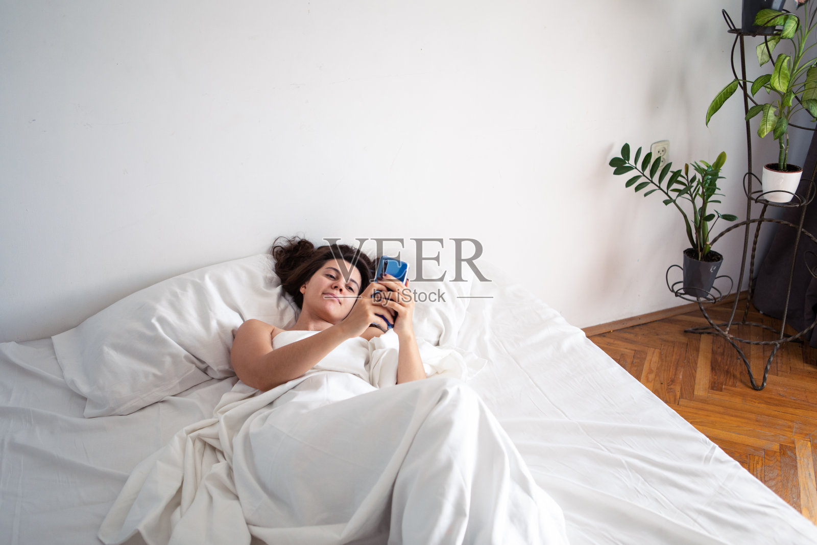 女人拿着智能手机躺在床上，铺着白床单照片摄影图片