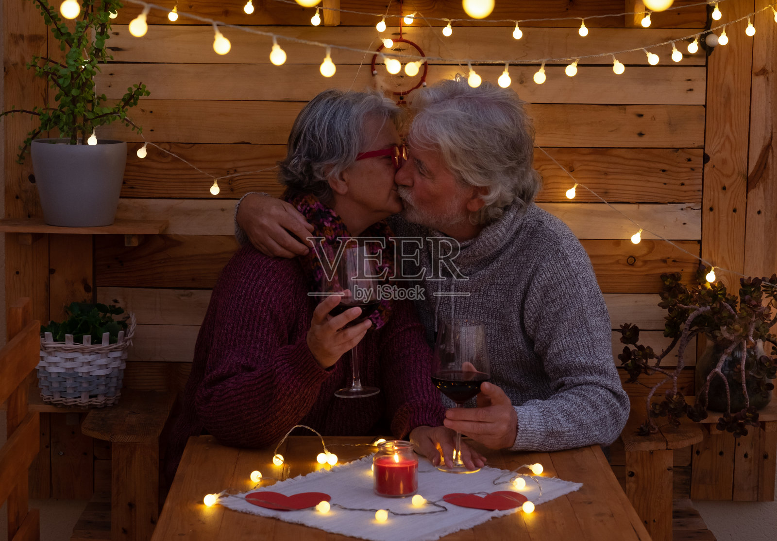 一个年长的男人和一个女人坐在户外一个浪漫的木制角落里温柔的亲吻。黄色的灯光。两杯红酒来庆祝一个积极的时刻照片摄影图片