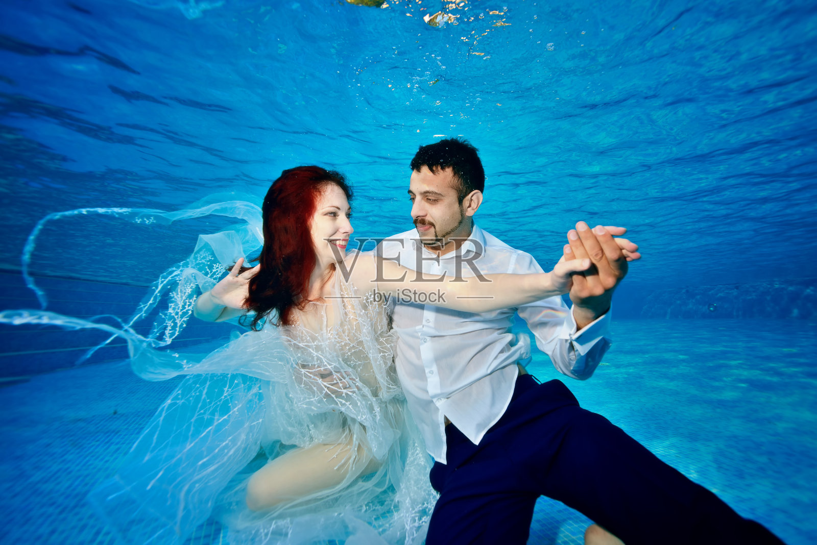 一对相爱的夫妇，一个女孩和一个混血男孩在一个阳光明媚的日子里穿着婚纱在室外游泳池游泳和玩耍。不同寻常的婚礼。特写镜头。横向。照片摄影图片