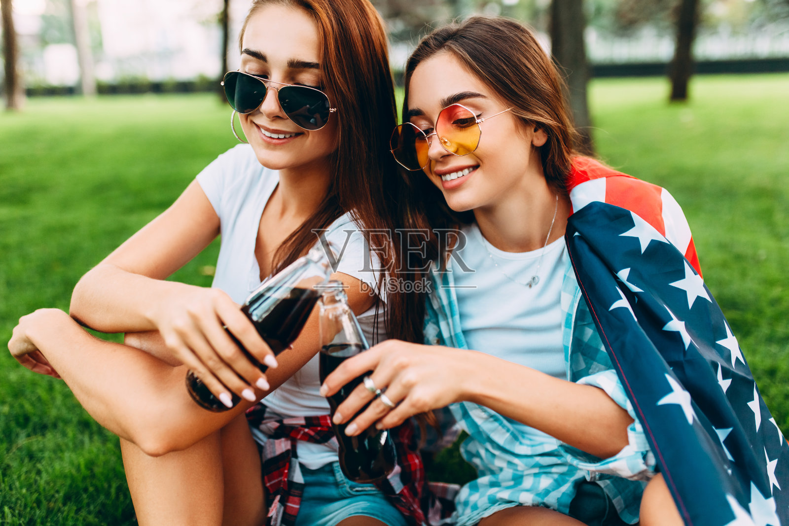 两个迷人的年轻女孩戴着太阳镜坐在公园的草坪上，旁边是美国国旗，喝着清爽的饮料照片摄影图片