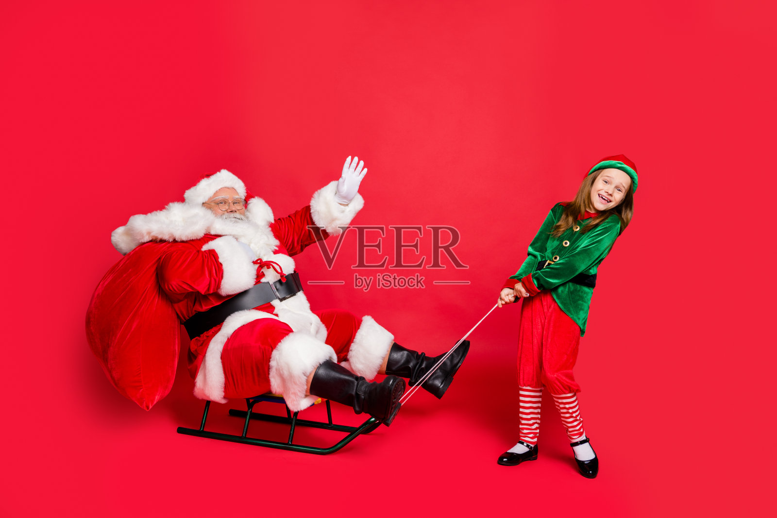 两个可爱的圣诞老人小精灵与红头发的灰头发挥舞着拉雪橇的袋子，戴着绿帽子，头戴眼镜，眼镜孤立在红色的背景照片摄影图片